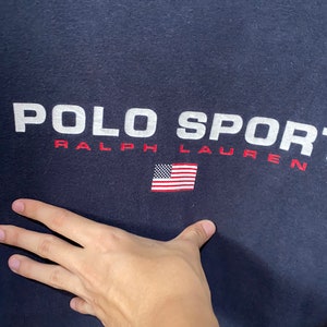 May include: A navy blue t-shirt with the text "POLO SPORT RALPH LAUREN" in white and red. A small American flag is embroidered on the shirt.