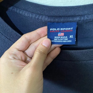 May include: A navy blue t-shirt with a white Polo Sport Ralph Lauren logo and the text "Polo Sport" in white. The tag says "Made in USA 100% Cotton" and "See Reverse for Care". The tag also has a size XL label.