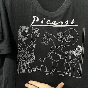 May include: Black t-shirt with a white line drawing of a Picasso painting. The painting depicts four figures, including a man playing a musical instrument. The text "Picasso" is printed in white at the top of the shirt.