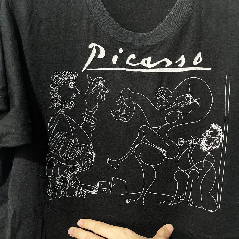 May include: Black t-shirt with a white line drawing of a Picasso painting. The painting depicts four figures, including a man playing a musical instrument. The text "Picasso" is printed in white at the top of the shirt.