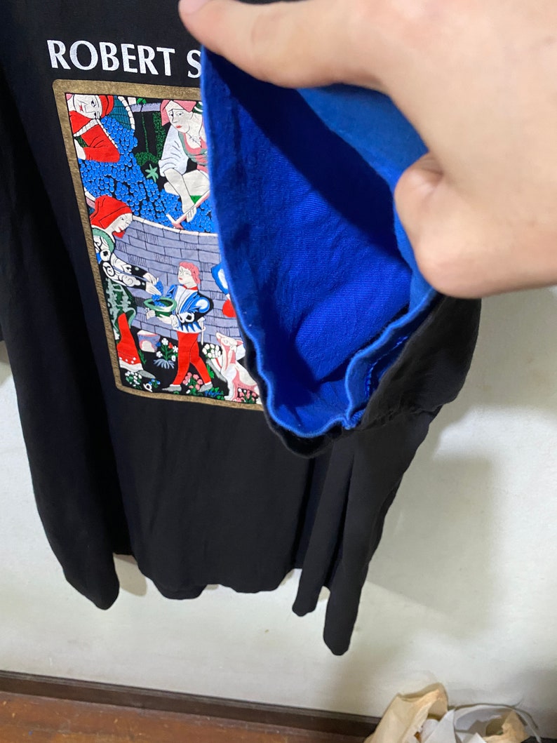 May include: Black t-shirt with a colorful illustration of people working in a vineyard. The illustration is framed in gold and has the text "ROBERT S" at the top. The t-shirt has a blue lining.