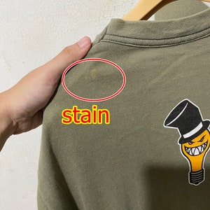 May include: A green t-shirt featuring a light bulb graphic wearing a top hat and a mischievous grin. The shirt has a small stain on the back.