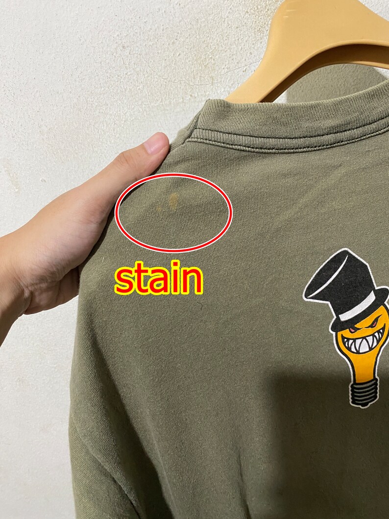 May include: A green t-shirt featuring a light bulb graphic wearing a top hat and a mischievous grin. The shirt has a small stain on the back.