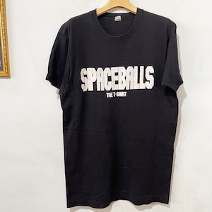 May include: Black t-shirt with the text "SPACEBALLS" in large, white, block letters. Below the main text is the smaller text "THE T-SHIRT". The shirt has short sleeves and a crew neck.
