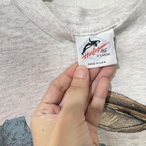 May include: Close-up of a gray t-shirt with a tag that reads "Harlequin NG X-Large Made in U.S.A." The tag features a black and white orca whale graphic. The shirt has a nature-themed graphic at the bottom.
