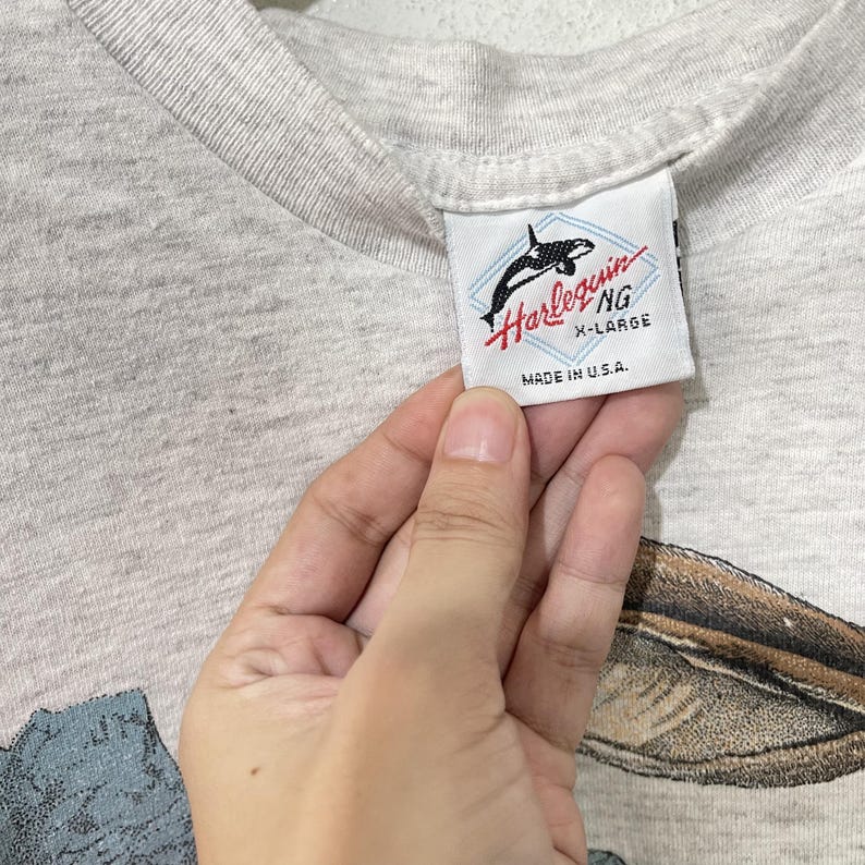May include: Close-up of a gray t-shirt with a tag that reads "Harlequin NG X-Large Made in U.S.A." The tag features a black and white orca whale graphic. The shirt has a nature-themed graphic at the bottom.