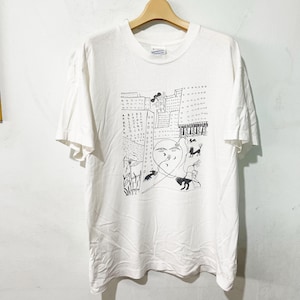 May include: White t-shirt with a black and white graphic design. The design features a face, buildings, and abstract figures. The shirt is hanging on a wooden hanger.
