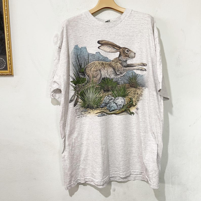 May include: A light gray t-shirt featuring a detailed illustration of a jackrabbit leaping across a desert landscape. The design includes desert plants, rocks, and a lizard. The shirt is hanging on a wooden hanger.