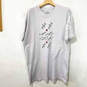 May include: A pale grey t-shirt with a crew neck and short sleeves. The shirt displays a colourful graphic design of interconnected circles and dots in shades of red, green, and yellow. The t-shirt is hanging on a wooden hanger.