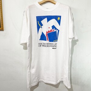 Op de afbeelding: Wit t-shirt met een blauw en wit grafisch ontwerp en de tekst "FOR THE MODERN ART OF PROGRAMMING" en het Microsoft-logo. De afbeelding bevat abstracte vormen en een gele ster.