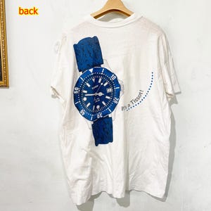 Puede incluir: Camiseta blanca con un gran gráfico azul y blanco de un reloj Tissot. El diseño del reloj incluye una correa azul y una esfera azul con números blancos. El texto "It's a Tissot!" está impreso en el lado derecho de la camiseta.