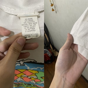 May include: White t-shirt with a colorful graphic print of a world map, mountains, and a seagull. The tag reads: RN #15490 50% Cotton 50% Polyester Machine Wash Warm Tumble Dry Low Do Not Bleach Do Not Iron If Decorated Catalog No. 514 One Size Fits All
