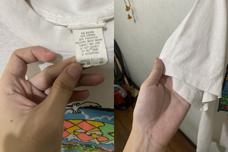 May include: White t-shirt with a colorful graphic print of a world map, mountains, and a seagull. The tag reads: RN #15490 50% Cotton 50% Polyester Machine Wash Warm Tumble Dry Low Do Not Bleach Do Not Iron If Decorated Catalog No. 514 One Size Fits All