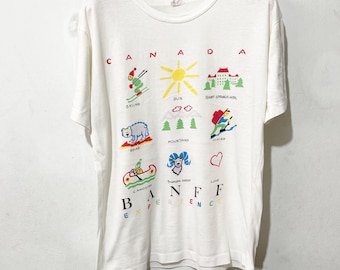 Könnte beinhalten: Weißes T-Shirt mit farbenfrohem Grafikdesign. Das Design zeigt das Wort "CANADA" und Illustrationen von Skifahren, einer Sonne, einem Hotel, einem Bären, Bergen, Wandern, einem Kanu, einem Dickhornschaf und den Worten "BANFF EXPERIENCE".