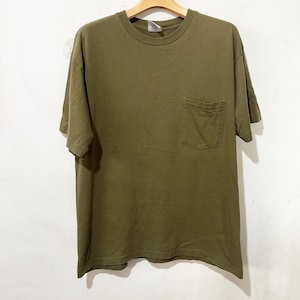 Pode incluir: Camiseta verde oliva com bolso no peito. A camiseta de manga curta está pendurada em um cabide de madeira. A camisa tem gola redonda e um ajuste folgado. O tecido parece ser uma mistura macia de algodão.