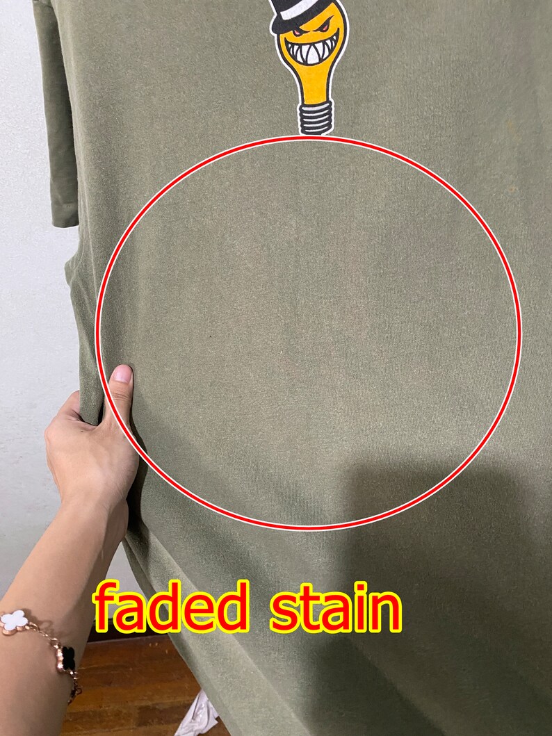 May include: A green t-shirt with a faded stain. The t-shirt features a cartoon lightbulb wearing a top hat and with a mischievous grin. The text "faded stain" is written in red below the lightbulb.