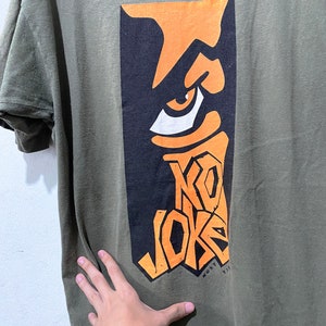 May include: A green t-shirt featuring a graphic print of a stylised eye and the text "KO JOKE" in orange and black. The text "MOST VIP" is printed in white below the graphic.