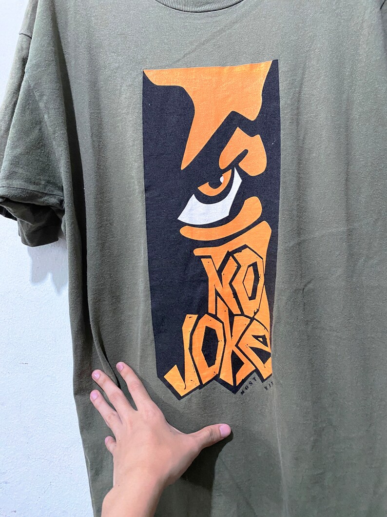May include: A green t-shirt featuring a graphic print of a stylised eye and the text "KO JOKE" in orange and black. The text "MOST VIP" is printed in white below the graphic.