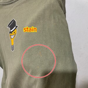 May include: A green t-shirt featuring a cartoon character wearing a top hat. There is a stain on the front of the shirt, circled in red.