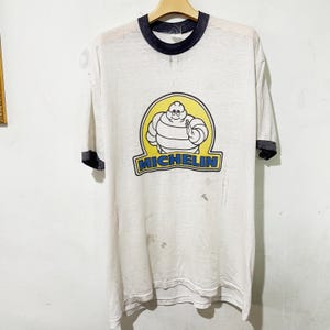 Puede incluir: Camiseta blanca vintage con cuello y mangas negras. La camiseta presenta el logotipo de Michelin Man en un óvalo amarillo con la palabra "MICHELIN" debajo. La camiseta está colgada de una percha de madera.