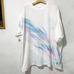 May include: A white short-sleeved t-shirt featuring a diagonal graphic print across the front. The design consists of a repeating hexagonal mesh pattern in light blue, pink, and yellow, with a small stylised dragon illustration on the upper right.