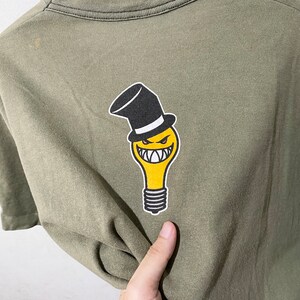 May include: A green t-shirt featuring a yellow lightbulb graphic wearing a black top hat. The lightbulb has a smiling face with sharp teeth.