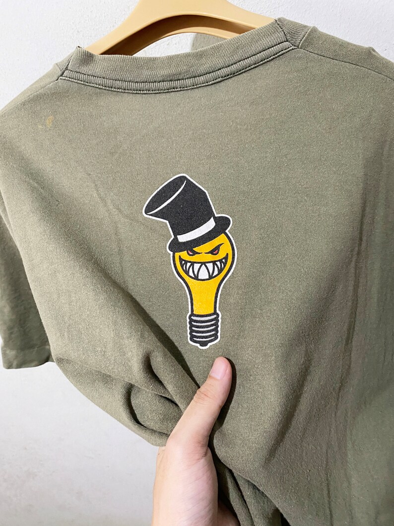 May include: A green t-shirt featuring a yellow lightbulb graphic wearing a black top hat. The lightbulb has a smiling face with sharp teeth.