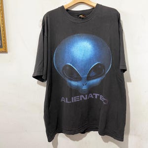 May include: Dark gray t-shirt featuring a large blue alien head graphic. The word "ALIENATED" is printed in a faded purple font below the alien. The shirt is hanging on a wooden hanger.