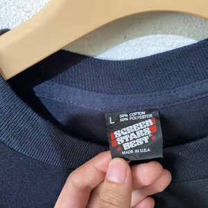 以下が含まれることがあります： 黒いTシャツにScreen Stars Bestのタグが付いています。タグは黒地に白文字で、赤い星が付いています。タグには「綿50％ ポリエステル50％」と「アメリカ製」と書いてあります。
