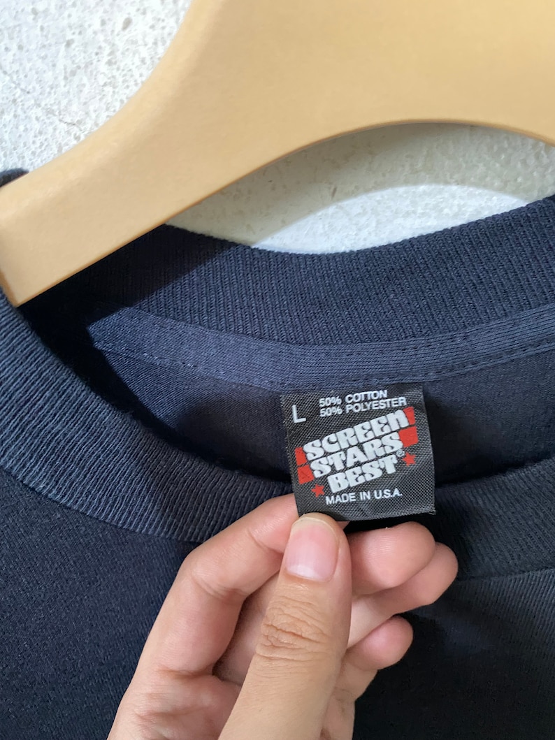 以下が含まれることがあります： 黒いTシャツにScreen Stars Bestのタグが付いています。タグは黒地に白文字で、赤い星が付いています。タグには「綿50％ ポリエステル50％」と「アメリカ製」と書いてあります。