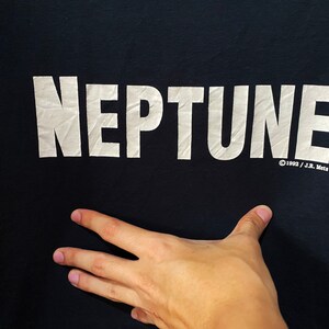 以下が含まれることがあります： 「NEPTUNE」と「&copy;1992 / J.R. Mets」という白い文字が書かれた黒いTシャツ。
