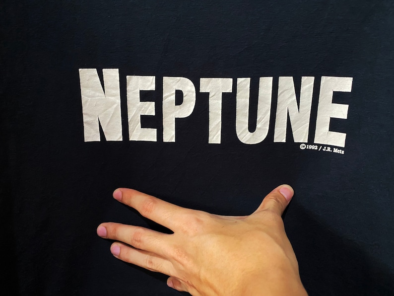 以下が含まれることがあります： 「NEPTUNE」と「&copy;1992 / J.R. Mets」という白い文字が書かれた黒いTシャツ。