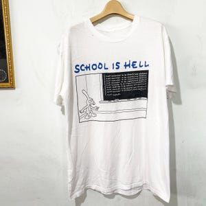 Op de afbeelding: Een wit T-shirt met korte mouwen en een grafische print met de blauwe tekst SCHOOL IS HELL boven een illustratie van een zwart schoolbord. De afbeelding toont een getekend konijn voor een schoolbord vol met de herhaalde zin I must remember to be cheerful and obedient.