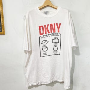 May include: White DKNY t-shirt with a red logo and graphic. The graphic includes the words "care contents" and icons for kisses, hugs, gifts, and trust. The shirt is hanging on a wooden hanger.