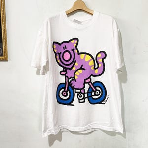 Könnte beinhalten: Weißes T-Shirt mit einer Cartoon-Katze auf einem Fahrrad. Die Katze ist lila mit gelben Punkten, und das Fahrrad ist blau und rosa. Das Design ist mittig platziert, mit einem einfachen, verspielten Stil.