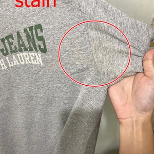 Puede incluir: Camiseta gris con una mancha en la manga. La camiseta tiene el texto "JEANS" y "RALPH LAUREN" impreso en la parte delantera.