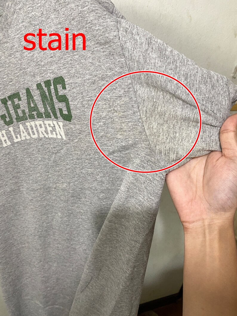 Puede incluir: Camiseta gris con una mancha en la manga. La camiseta tiene el texto "JEANS" y "RALPH LAUREN" impreso en la parte delantera.
