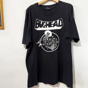 May include: Black t-shirt with the word "BUCHEAD" in a white rectangular box. Below the text is a white graphic of a person playing a musical instrument inside a circle. The t-shirt is hanging on a wooden hanger.