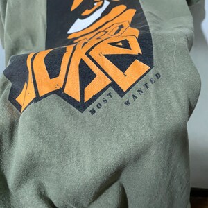 May include: Green t-shirt with a black and orange graphic of a face with the text "MOST WANTED" underneath.