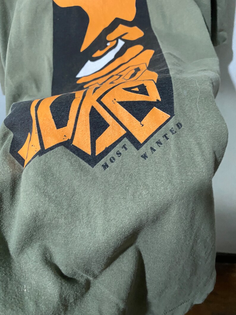 May include: Green t-shirt with a black and orange graphic of a face with the text "MOST WANTED" underneath.