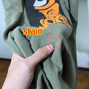 May include: A green t-shirt with a graphic print of an orange eye and the word "stain" written on the shirt. There is a small white stain on the shirt.