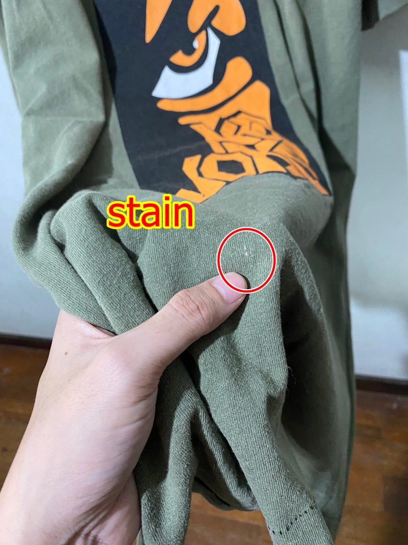 May include: A green t-shirt with a graphic print of an orange eye and the word "stain" written on the shirt. There is a small white stain on the shirt.