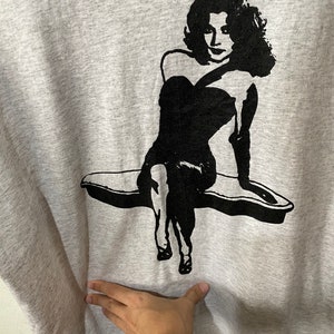 Puede incluir: Una sudadera gris con un gr&aacute;fico negro de una mujer con un vestido sin tirantes. La mujer est&aacute; sentada en una superficie curva. El gr&aacute;fico es un dise&ntilde;o simple y audaz.