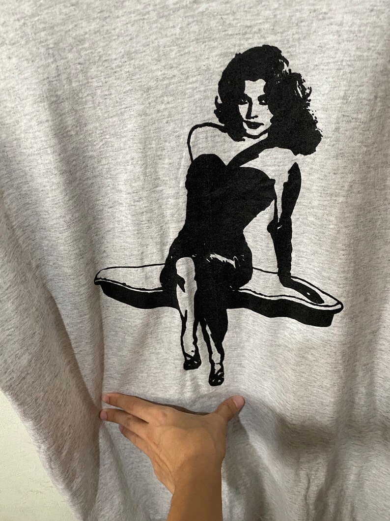 Puede incluir: Una sudadera gris con un gr&aacute;fico negro de una mujer con un vestido sin tirantes. La mujer est&aacute; sentada en una superficie curva. El gr&aacute;fico es un dise&ntilde;o simple y audaz.