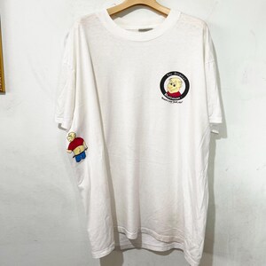 May include: White t-shirt featuring a cartoon character with the text "The Original Buttman". The shirt has a small graphic on the sleeve and a larger circular graphic on the chest. The shirt is hanging on a wooden hanger.