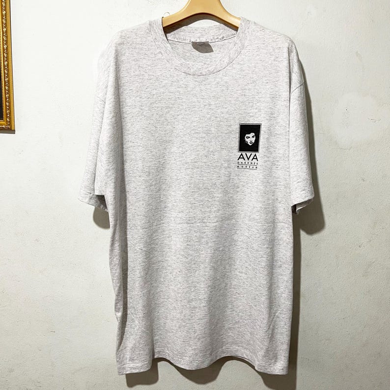 Puede incluir: Una camiseta gris claro con cuello redondo y mangas cortas. La camiseta presenta un gr&aacute;fico cuadrado negro en la parte superior izquierda del pecho con una cara estilizada y el texto "AVA CALIFORNIA MOVIES" debajo. Cuelga de una percha de madera.