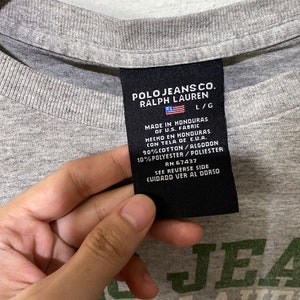 Puede incluir: Una camiseta gris con una etiqueta negra que dice "Polo Jeans Co. Ralph Lauren L/G Hecho en Honduras de tela de EE. UU. Hecho en Honduras con tela de E.U.A. 90% Algod&oacute;n/Algod&oacute;n 10% Poli&eacute;ster/Poli&eacute;ster RN 67437 Ver el reverso Cuidado Ver al Dorso". La camiseta tambi&eacute;n tiene un logotipo verde y blanco que dice "Polo Jeans Co. Ralph Lauren".