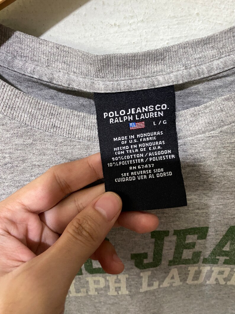 Puede incluir: Una camiseta gris con una etiqueta negra que dice "Polo Jeans Co. Ralph Lauren L/G Hecho en Honduras de tela de EE. UU. Hecho en Honduras con tela de E.U.A. 90% Algod&oacute;n/Algod&oacute;n 10% Poli&eacute;ster/Poli&eacute;ster RN 67437 Ver el reverso Cuidado Ver al Dorso". La camiseta tambi&eacute;n tiene un logotipo verde y blanco que dice "Polo Jeans Co. Ralph Lauren".