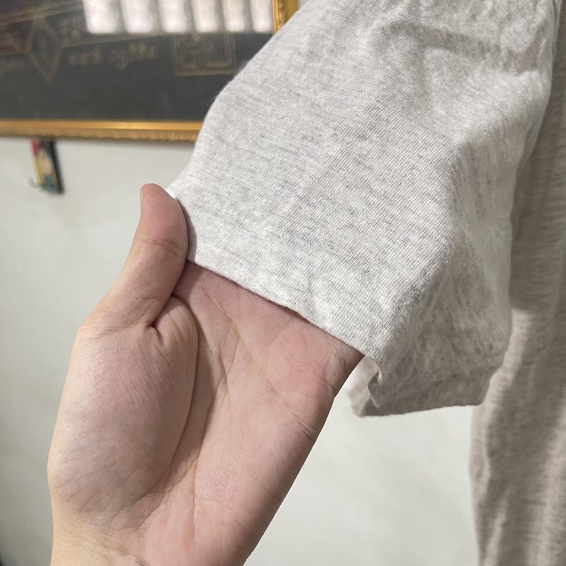 May include: Close-up of a light gray short-sleeved shirt. The fabric appears to be a soft, heathered material. The sleeve is visible, with a folded hem. The shirt is held by a hand.