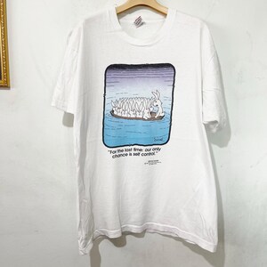 May include: White t-shirt featuring a cartoon illustration of rabbits in a boat. The image includes the text: "For the last time: our only chance is self control." The shirt is hanging on a wooden hanger.
